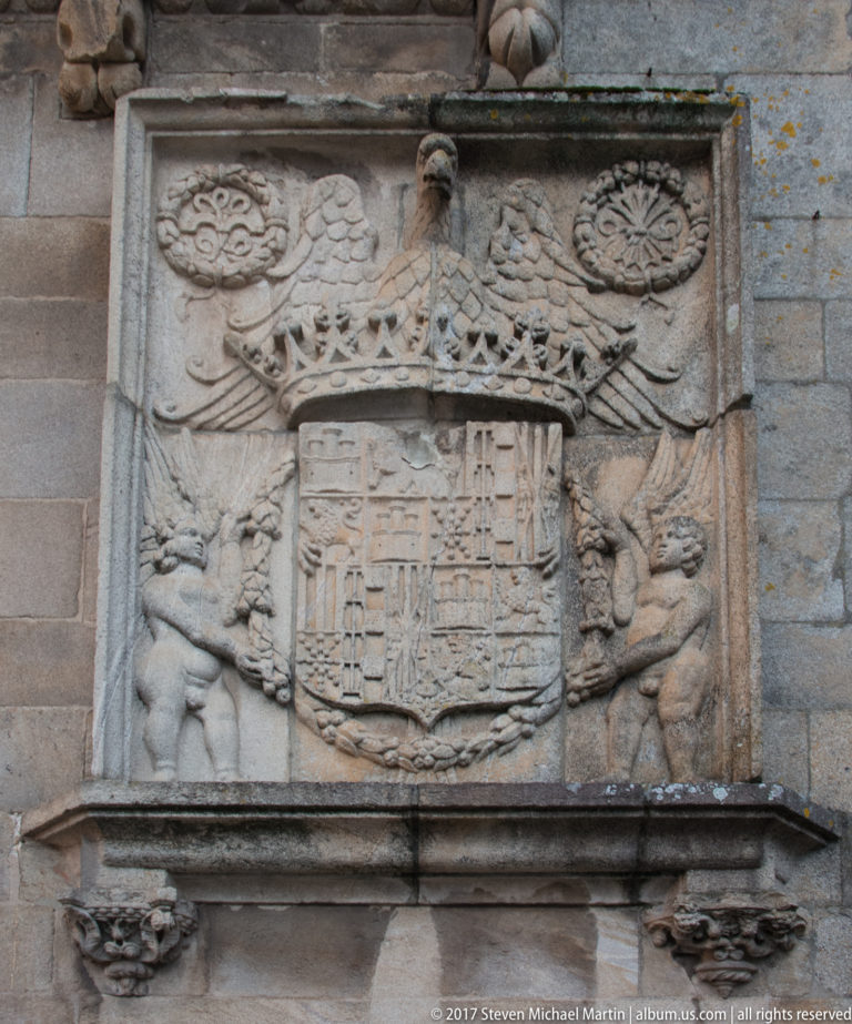 Weathered stone heraldic panel with a quartered shield, winged forms, carved supporters, and ornamental motifs.