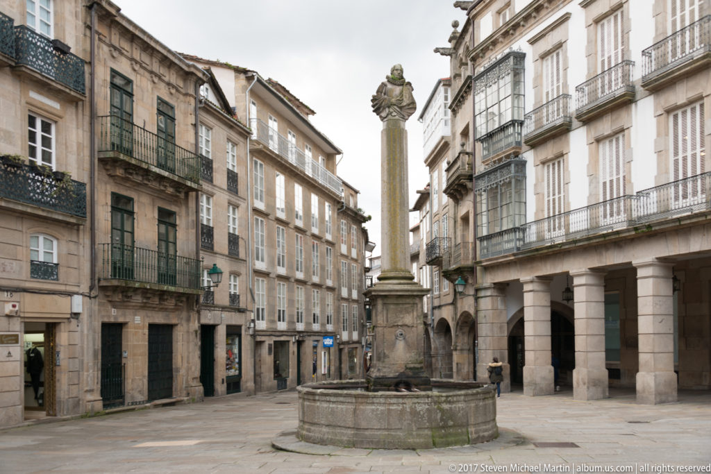 Stone column and circular fountain basin in a small square lined with multi-story buildings and iron balconies.
