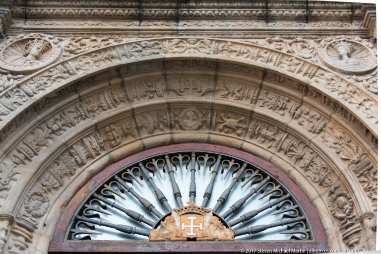 Close view of carved stone archivolts above a radiating metal fanlight centered on a small cross emblem.