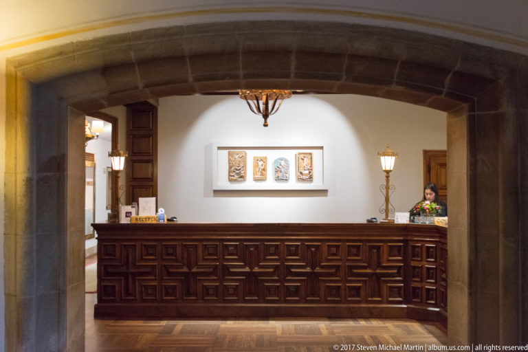 Dark carved-wood reception desk beneath a stone arch, with wall plaques, lanterns, and a staff member behind the counter.