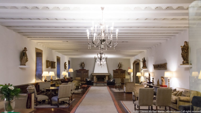 Long white lounge with chandeliers, ceiling beams, sculptures, upholstered chairs, lamps, rugs, and a fireplace at the far end.