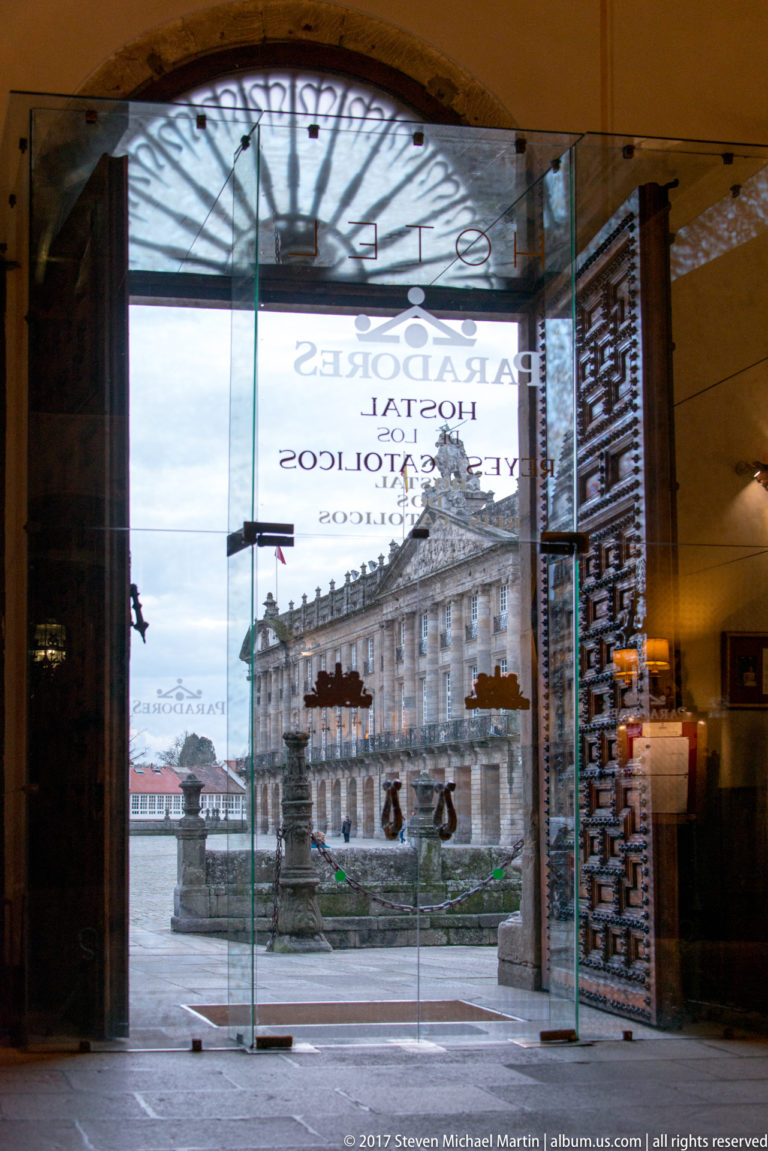 Glass hotel entrance with reversed “Paradores” and “Hostal Reyes Católicos” lettering, opening toward a stone square and historic buildings.