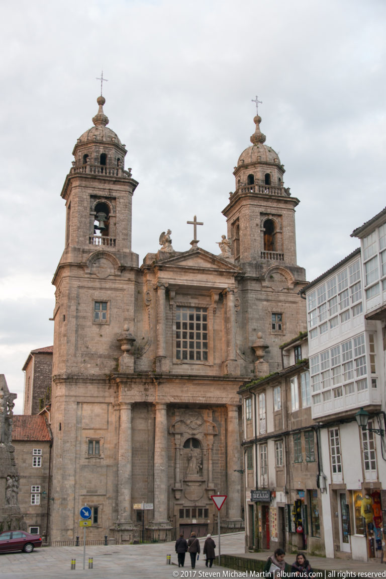 Frontal view of a granite church façade with twin bell towers, central window, carved portal, and neighboring city buildings.