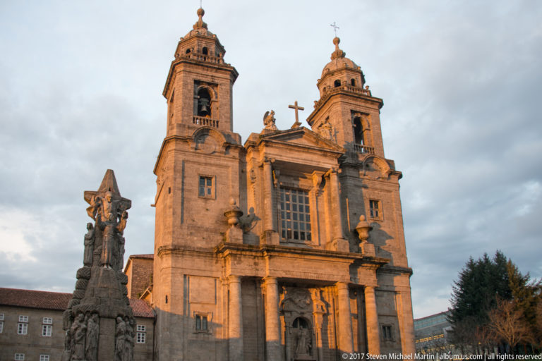 Low-angle evening view of a twin-towered stone church with a tall sculptural monument standing in the foreground.