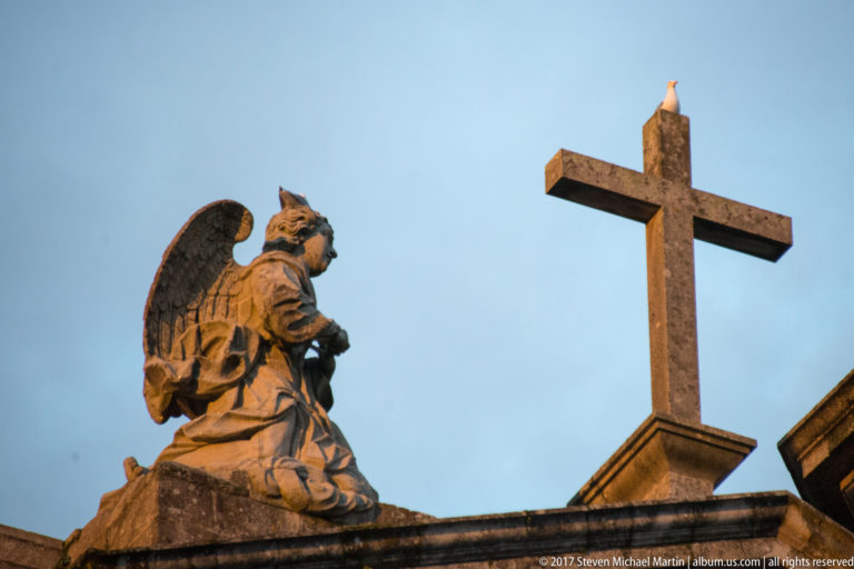 Kneeling winged stone figure on a roofline beside a large stone cross against a blue sky.