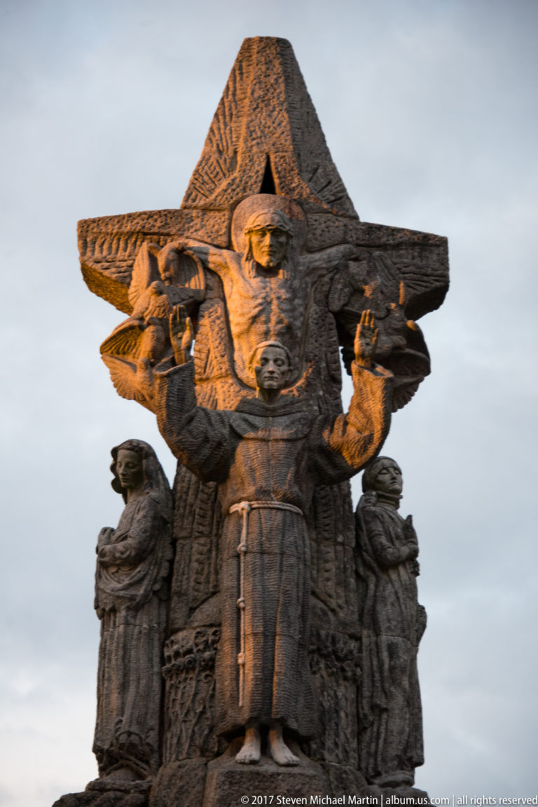 Large stone pilgrim figure in a broad hat and cloak, with smaller carved figures grouped around the base.