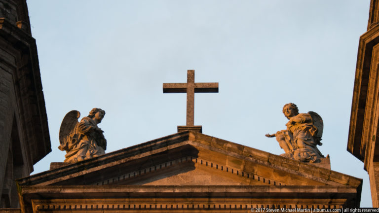 Stone cross centered on a triangular pediment, flanked by two carved religious figures against the sky.