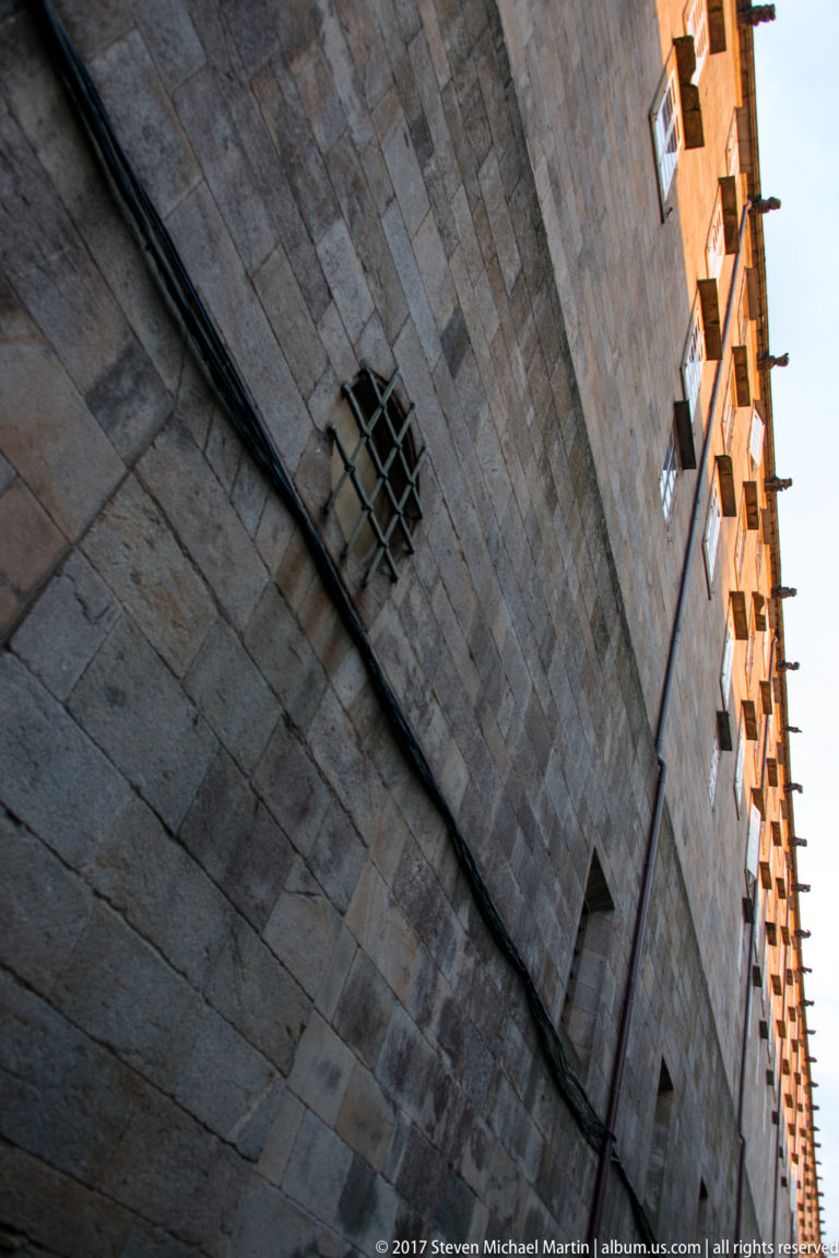 Oblique upward view of a tall granite wall with a small barred window and a narrow building edge beside it.