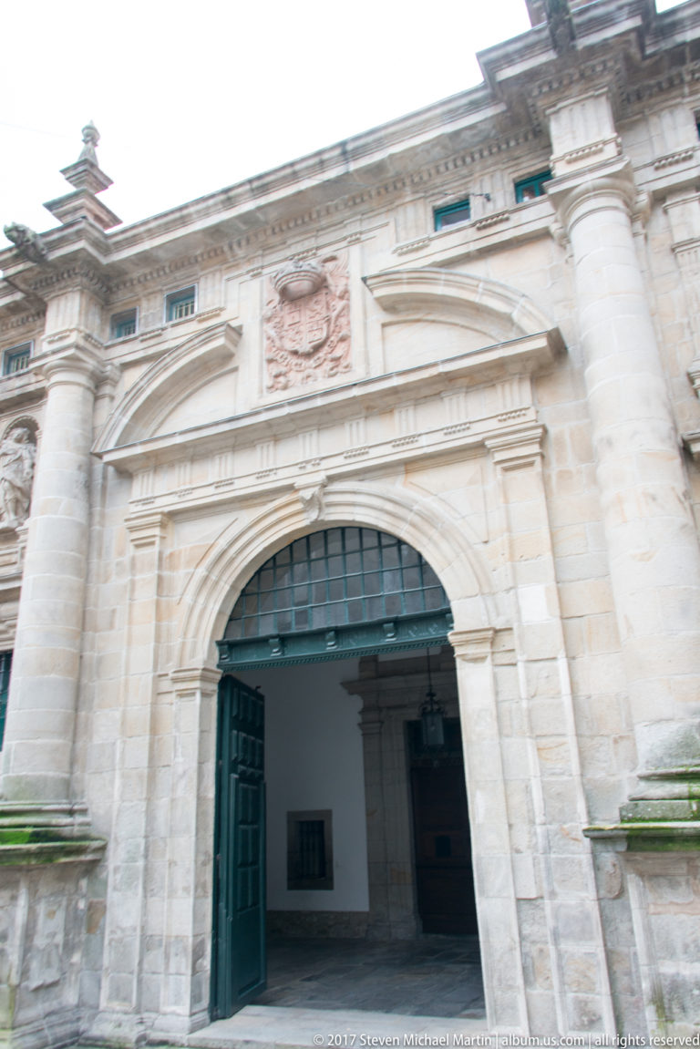 Large semicircular stone gateway with open blue-green doors, classical pilasters, heraldic relief, and a shadowed passage beyond.