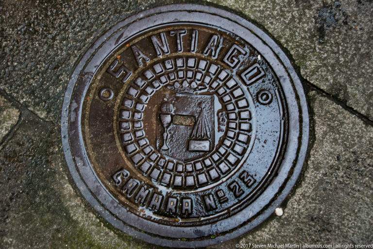 Round metal utility cover marked “SANTIAGO” with a central emblem, set into wet stone paving.