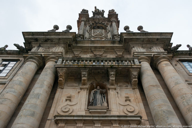 Low-angle detail of a monumental stone entrance with large columns, a niche statue, balcony, and heraldic crest above.