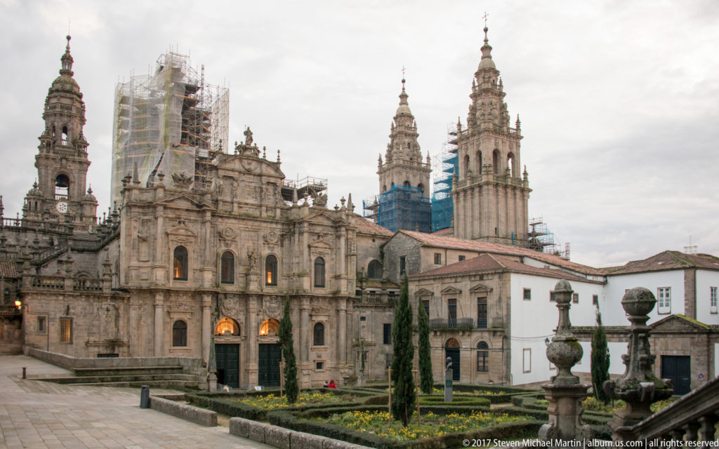 Santiago Cathedral towers and adjoining stone buildings seen beyond a formal garden, with scaffolding on one tower.