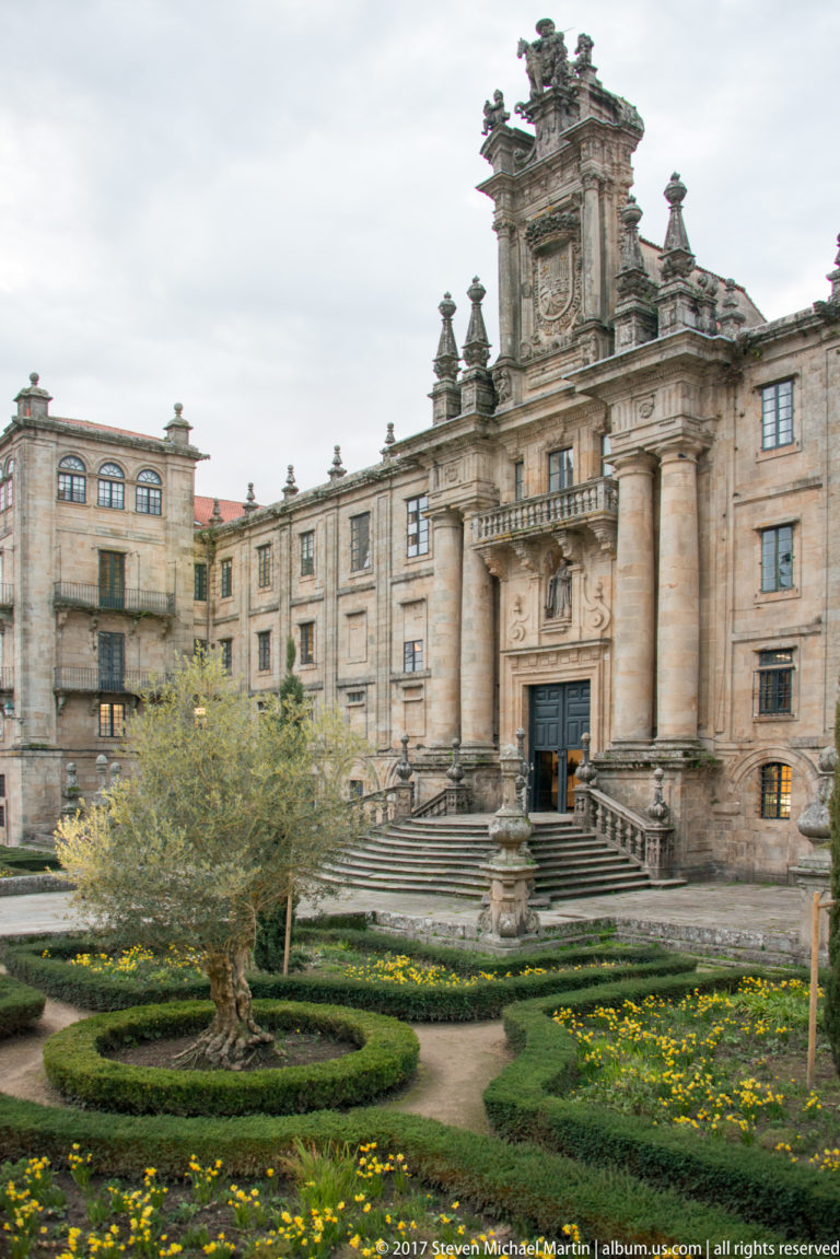 Formal hedge and flower garden in front of a grand granite building with a central staircase and columned entrance.