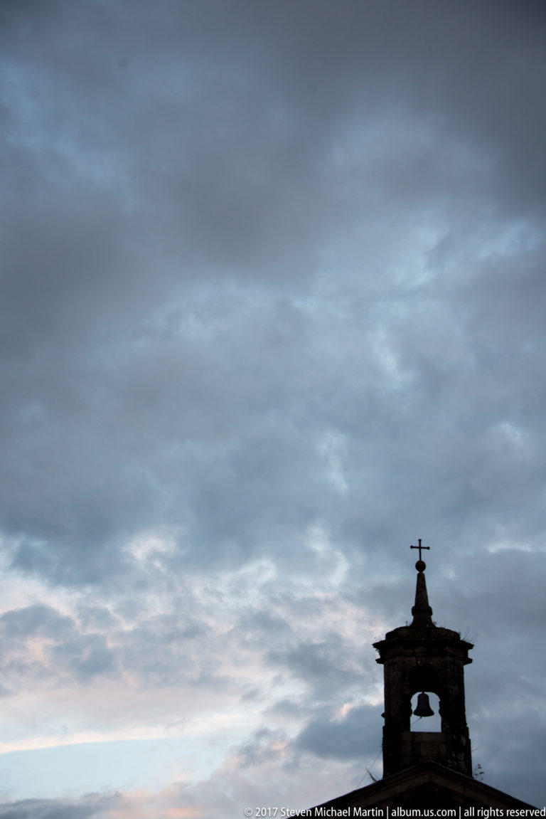 Small bell tower and cross silhouetted along the lower edge of a broad, dark cloud-filled evening sky.