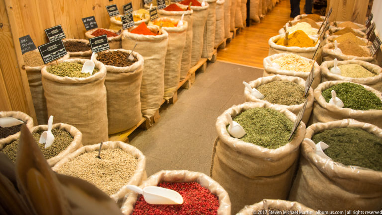 Rows of open burlap sacks filled with herbs, seeds, spices, and dried goods, with scoops and small labels.