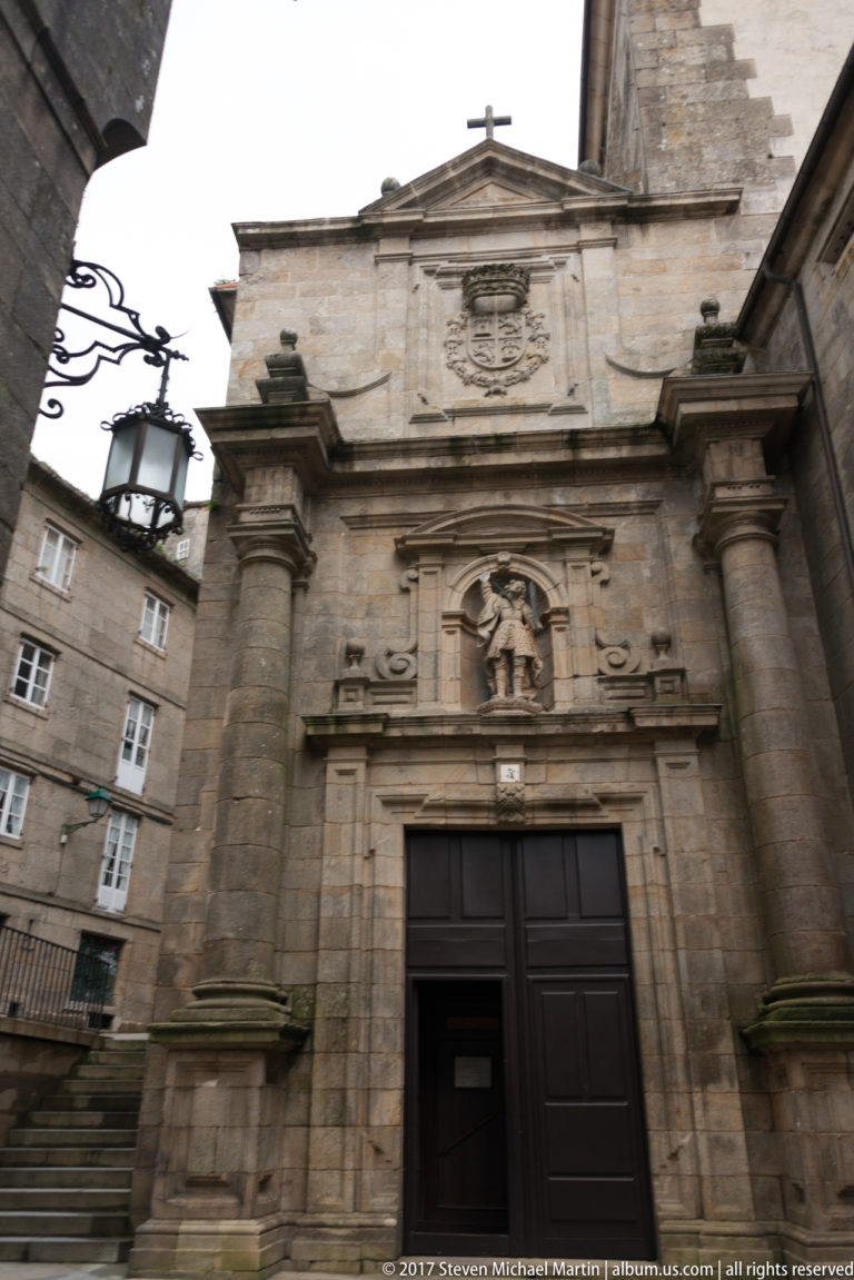 Stone church façade with dark wooden doors, paired pilasters, a saint statue in a niche, triangular pediment, and cross.