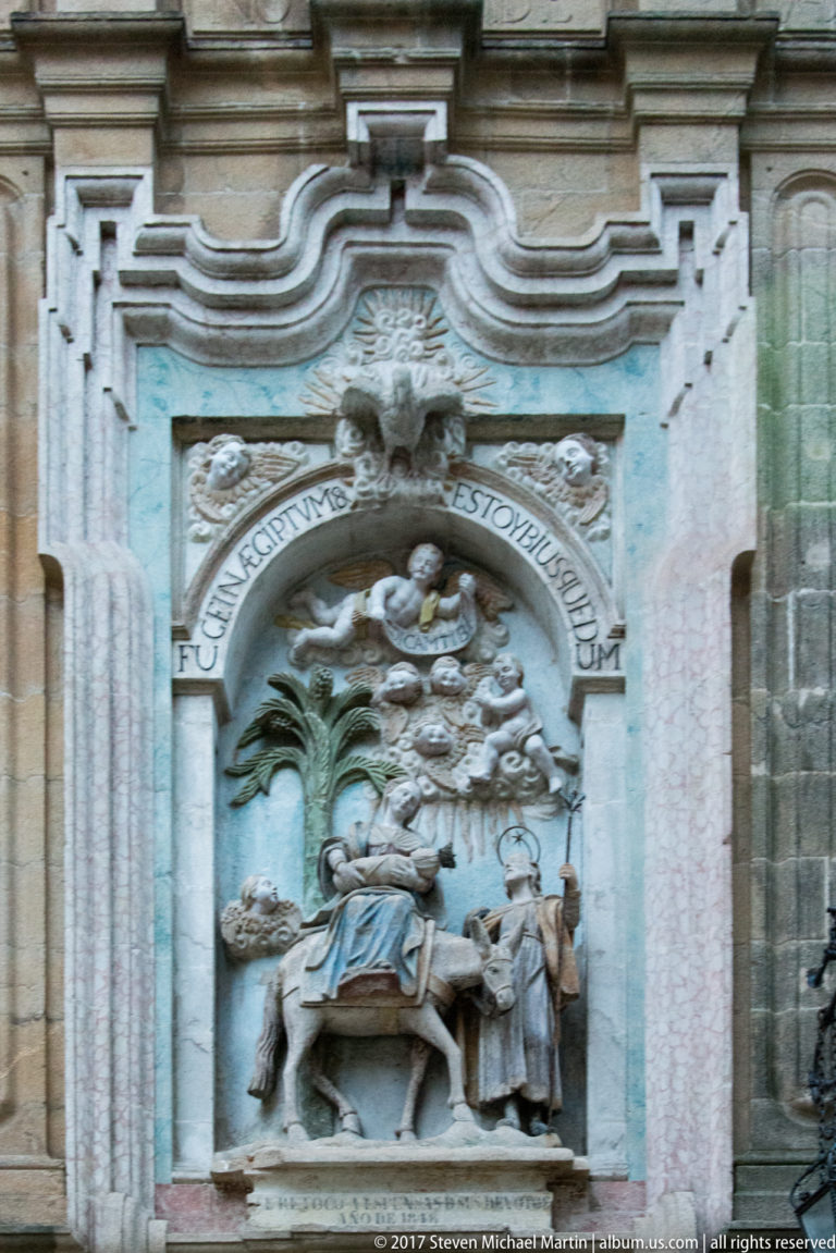 Ornate stone relief with seated and standing religious figures beneath a curved Latin inscription and decorative architectural framing.