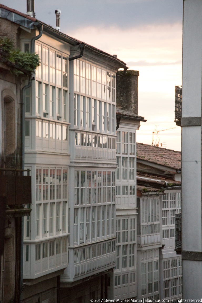 White multi-story glazed galleries on historic buildings, viewed obliquely at dusk above lower rooftops.