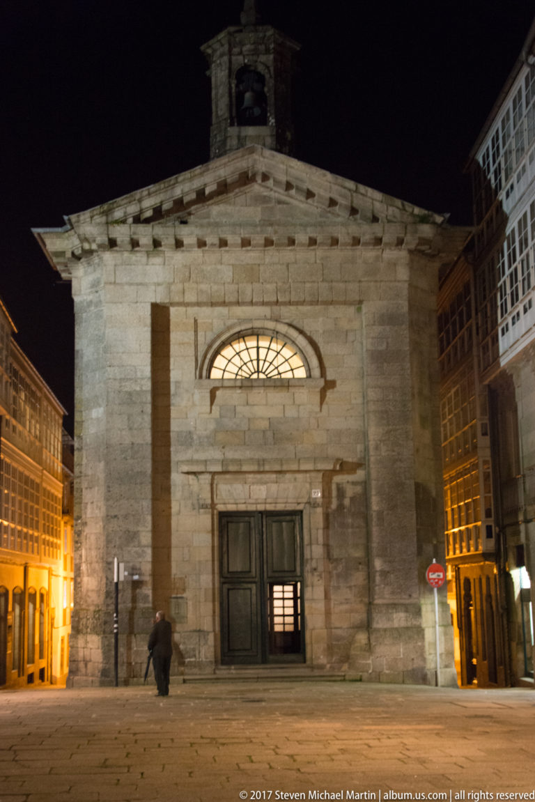 Small granite church façade at night with double doors, semicircular fanlight, bellcote, and taller glazed buildings around it.