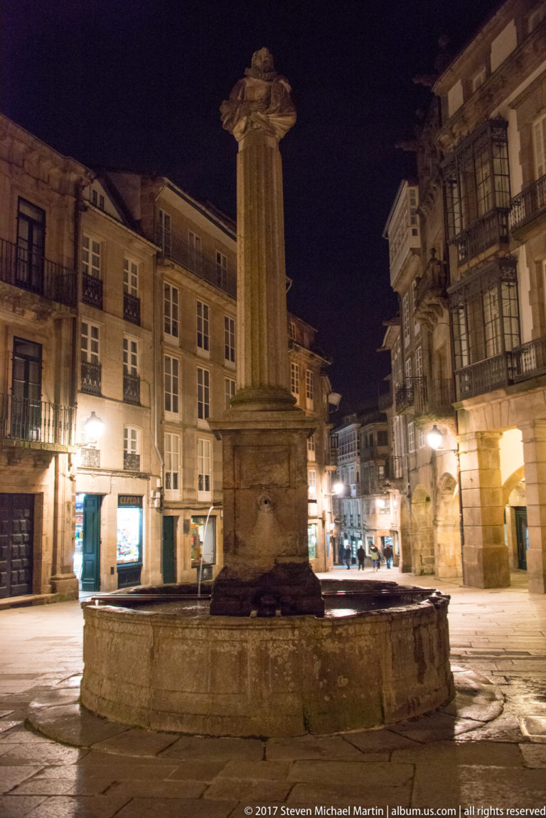 Tall stone column fountain in a historic square at night, surrounded by illuminated multi-story façades and balconies.