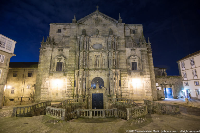 Illuminated granite church façade at night with sculpted niches, oval window, central doorway, and broad balustraded stairs.
