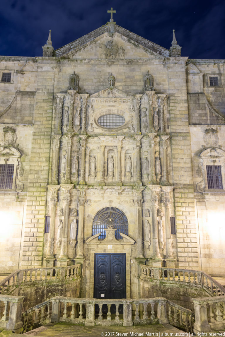 Close nighttime view of a sculpted granite church façade with niches, figures, arched opening, dark doors, and foreground balustrade.