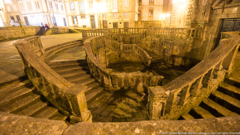 High-angle night view down into a symmetrical curving stone staircase with heavy balustrades and a rounded central landing.