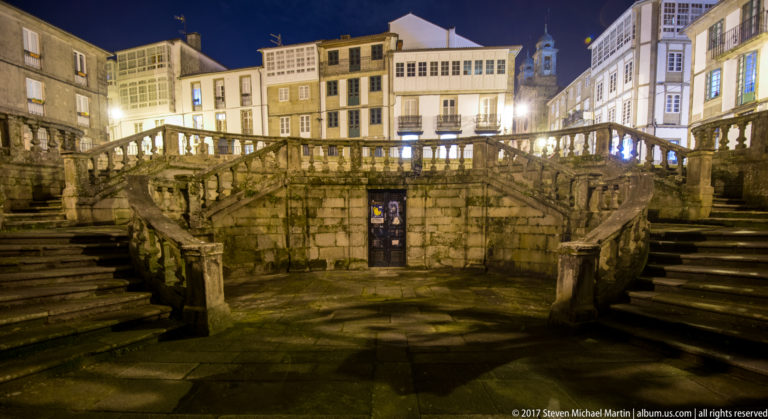 Frontal night view of a broad stone staircase with side flights, balustraded upper landing, central doorway, and lit buildings beyond.