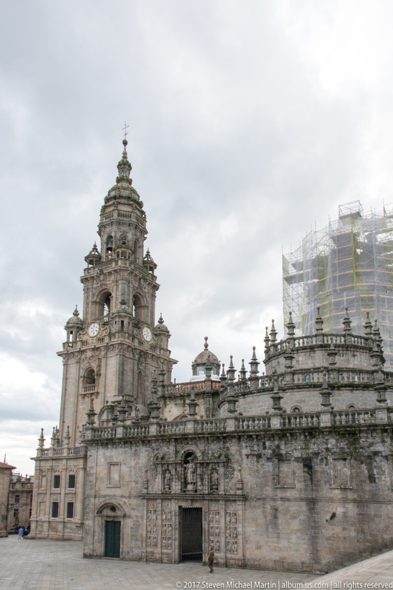 Wide cathedral side elevation with tower, sculpted portals, balustrade, and restoration scaffolding at the right.