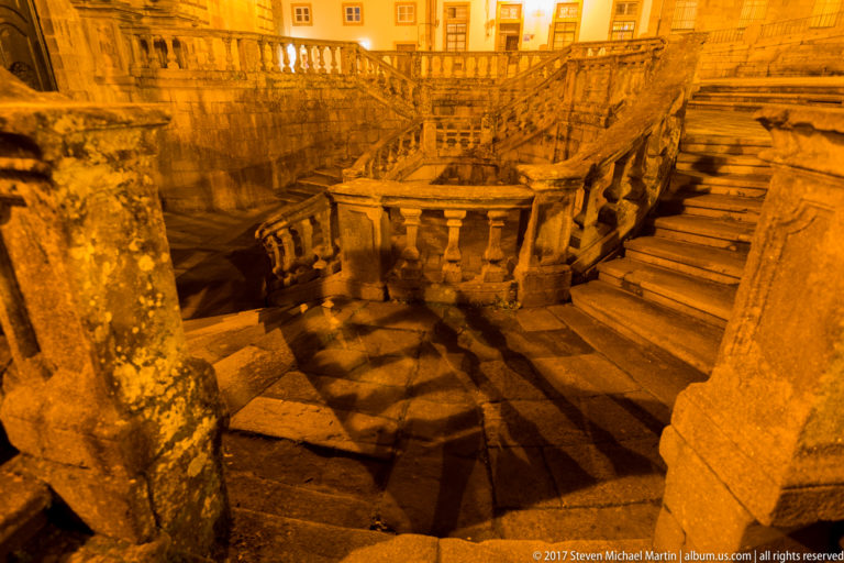 Warm-lit nighttime close view of curving granite steps and heavy stone balustrades crossing at multiple levels.