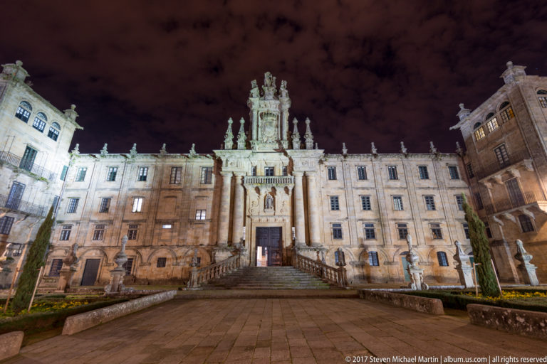 Illuminated Baroque façade of San Martiño Pinario with broad steps, central portal, columns, and rooftop sculpture.