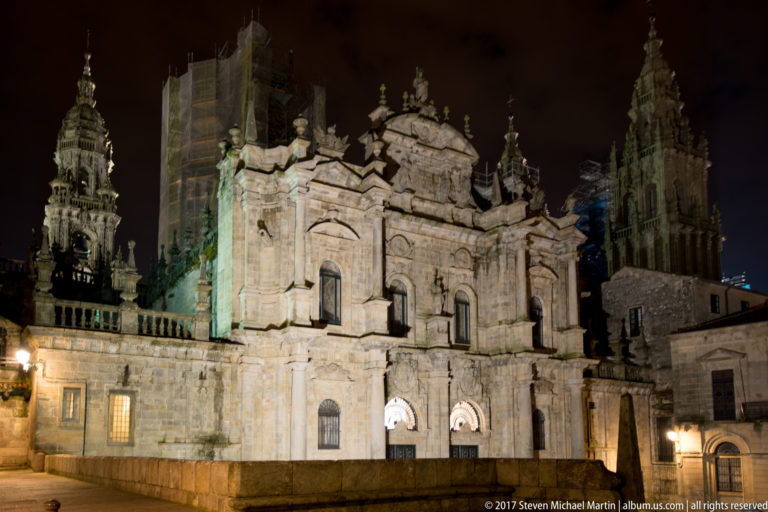Illuminated ornate stone cathedral elevation with arched doorways and towers rising behind it at night.