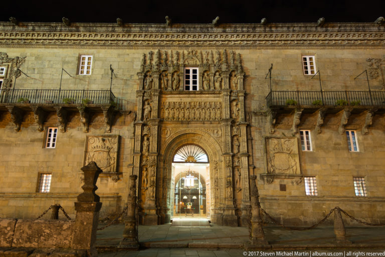 Ornate carved stone portal of the Hostal dos Reis Católicos illuminated at night on Praza do Obradoiro.