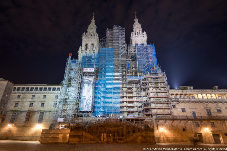 Twin cathedral towers and central façade covered by scaffolding and blue protective netting at night.