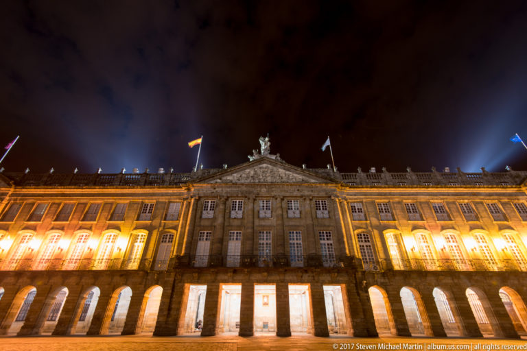 Illuminated Pazo de Raxoi with long ground-floor arcade, central pediment, and rooftop sculpture facing Praza do Obradoiro.