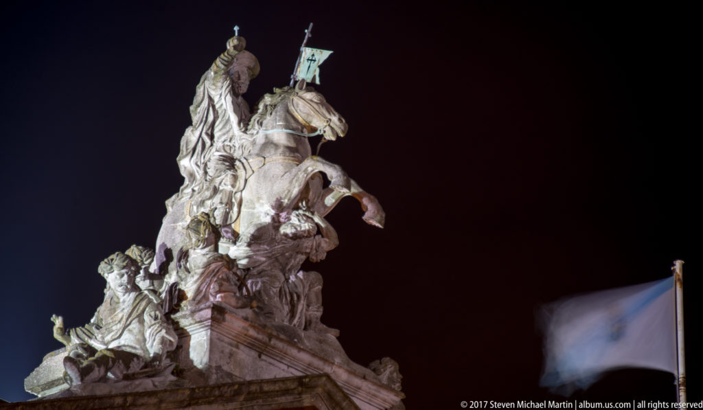 Illuminated stone equestrian sculpture of Saint James on a rearing horse, holding a standard against the night sky.
