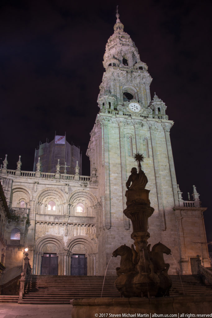 Dark sculptural fountain in front of an illuminated cathedral clock tower and entrance at night.
