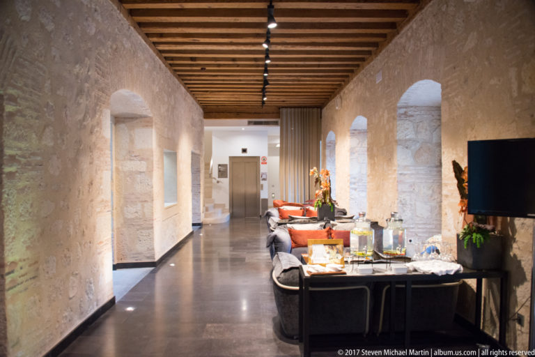 Long stone corridor with timber ceiling, arched openings, polished floor, lounge seating, dining tables, and modern lighting.