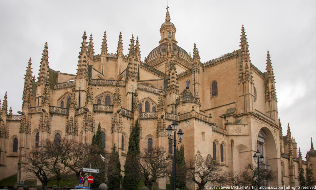 Rear and side exterior of Segovia Cathedral with pinnacles, buttresses, traceried windows, chapels, and dome under a cloudy sky.