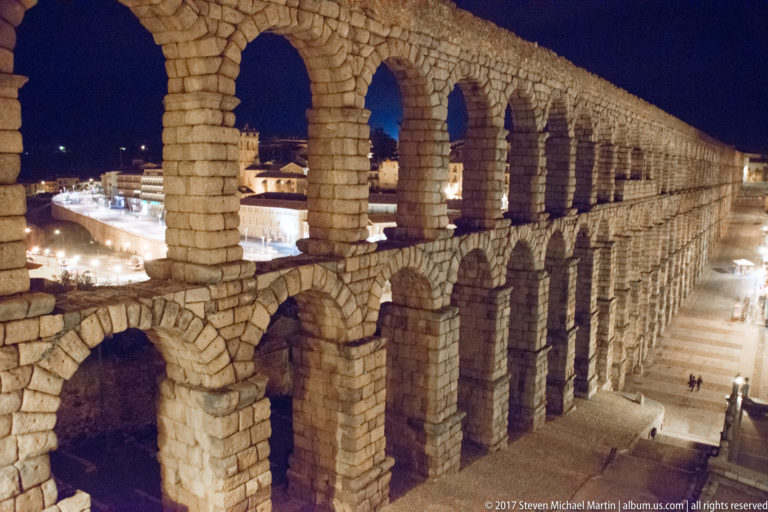 Oblique elevated night view along Segovia’s aqueduct, with stacked granite arches receding in perspective toward city lights.