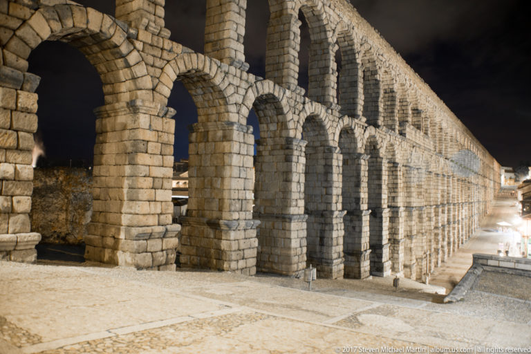 Ground-level night view beside Segovia’s Roman aqueduct, with huge granite piers and arches receding along a paved pedestrian space.