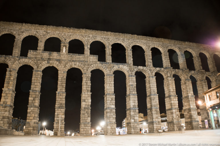Wide frontal night view of Segovia’s double-tiered Roman aqueduct, with tall granite arches rising above streetlights and small figures.