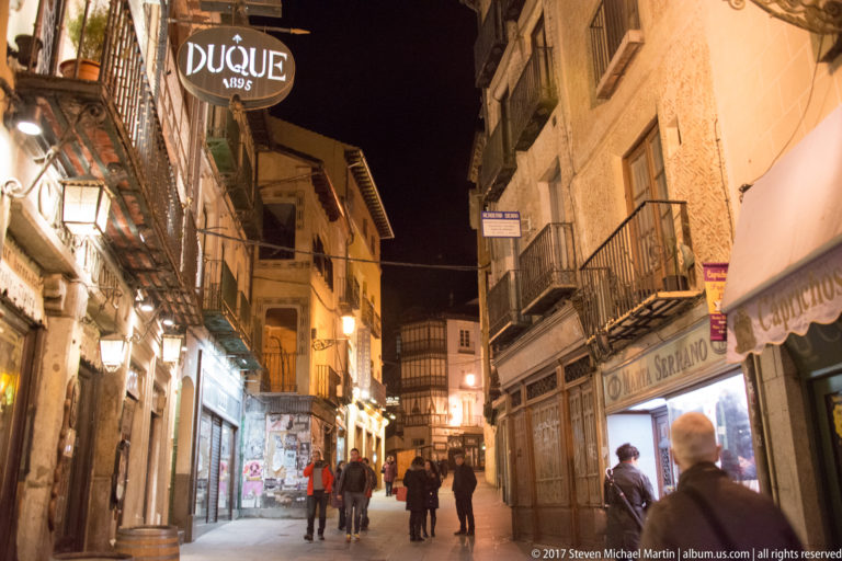 Night pedestrian street with iron balconies, lit shopfronts, hanging signs, and small groups walking along the narrow lane.