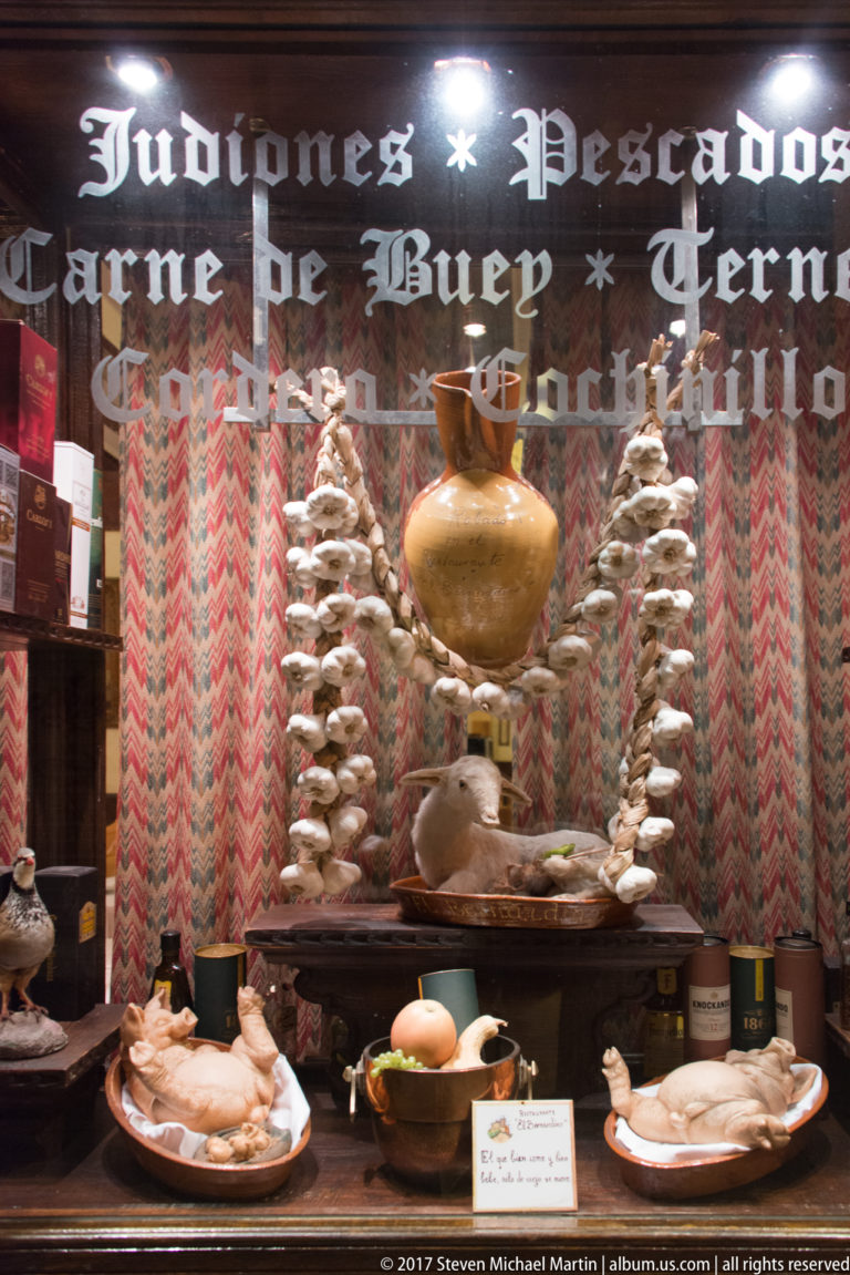 Restaurant window display with plates of meat, a large earthenware vessel, repeated decorative food forms, and painted Spanish menu lettering.