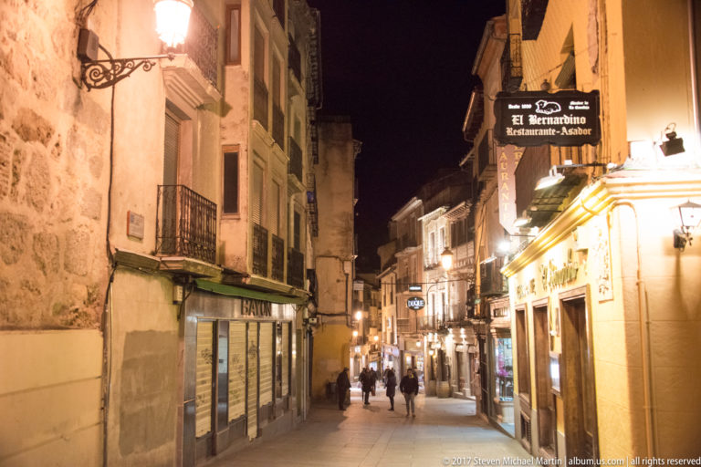 Night view down a gently descending narrow street with historic facades, balconies, restaurant signs, and pedestrians walking away.