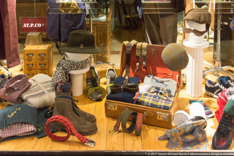Shop window filled with hats, scarves, belts, gloves, shoes, folded textiles, and leather accessories arranged around mannequin heads.