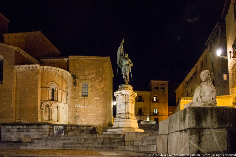 Night square with an equestrian statue on a tall pedestal, historic masonry buildings, and a seated stone figure in the foreground.