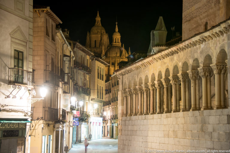 Night street beside a long Romanesque arcade of semicircular arches and paired columns, with church towers rising in the distance.