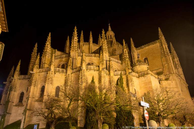 Illuminated exterior of Segovia Cathedral at night, showing the apse, flying buttresses, pinnacles, and bare trees in the foreground.
