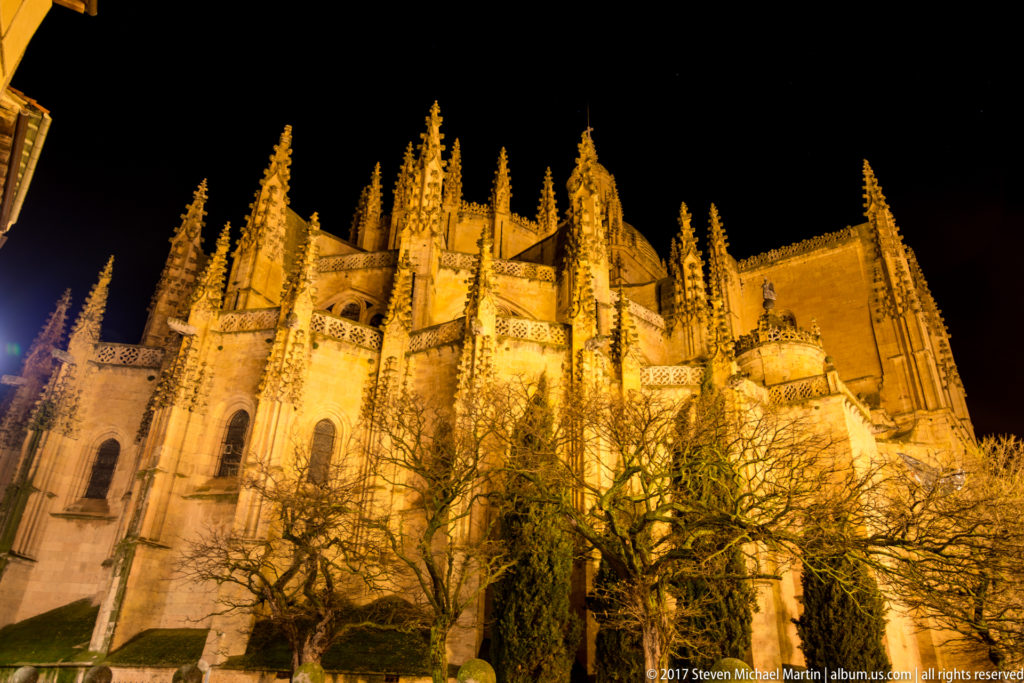 Wide night view of Segovia Cathedral’s illuminated side elevation, with buttresses and pinnacles rising behind leafless trees.